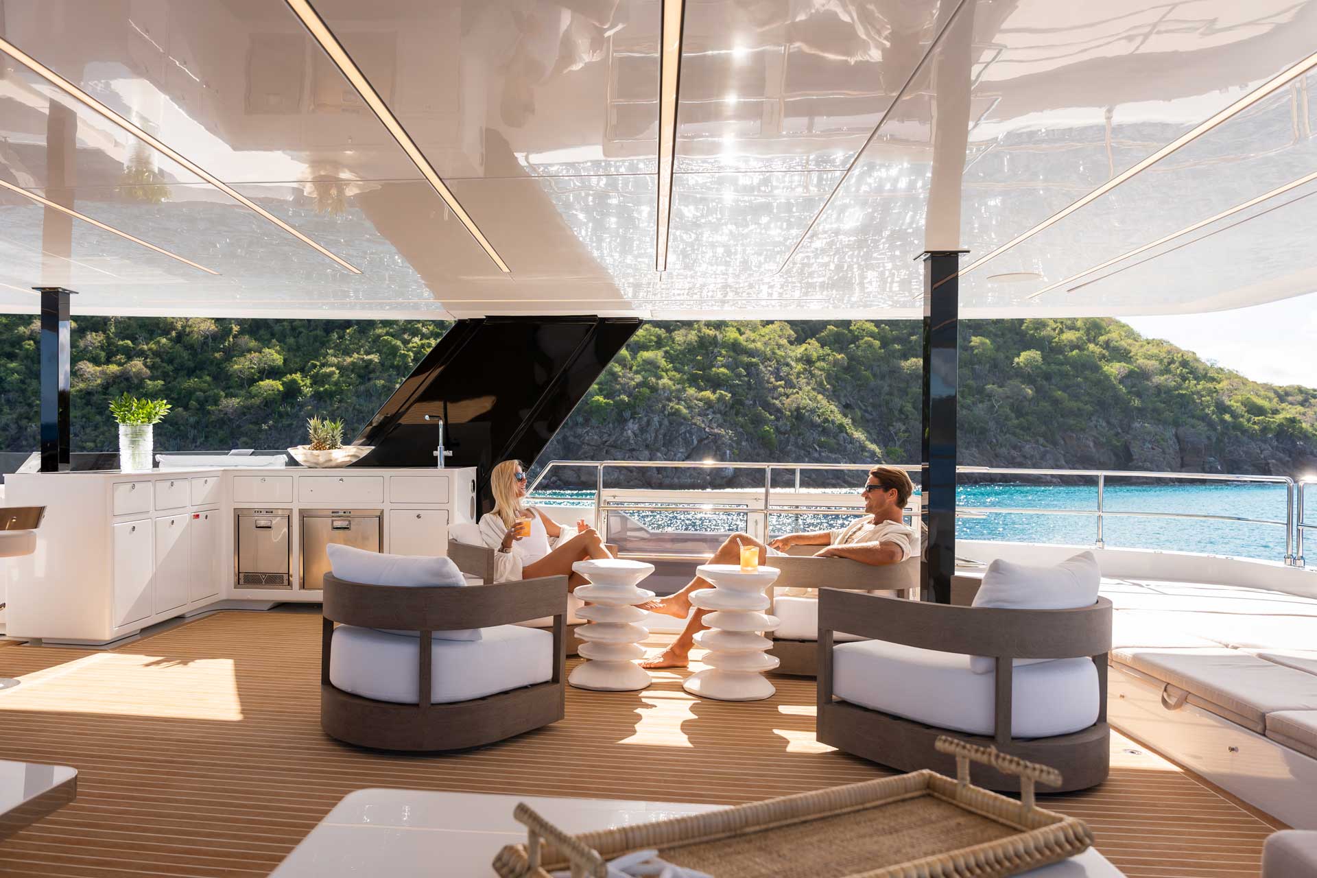 DSC04810 Two people relaxing on the deck of a Sunreef Yachts luxury catamaran with a view of the ocean and a lush coastline in the background.