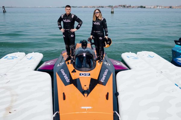 teamrafaXsunreef-web-4 Two people wearing Sunreef Yachts-branded race suits are standing next to a Team Rafa racing boat, with water around and the sky slightly overcast.